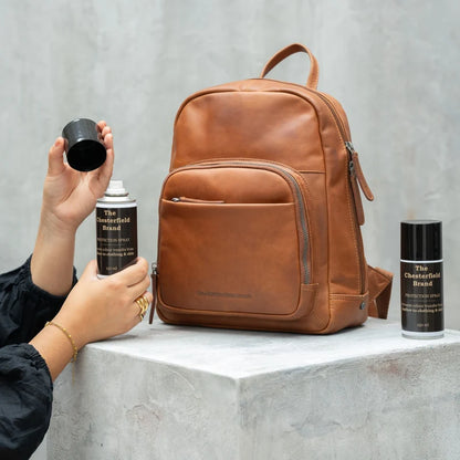 Chesterfield Colour Fix Schutzspray in Anwendung – Hand sprüht auf cognacfarbenen Lederrucksack, zweite Dose daneben