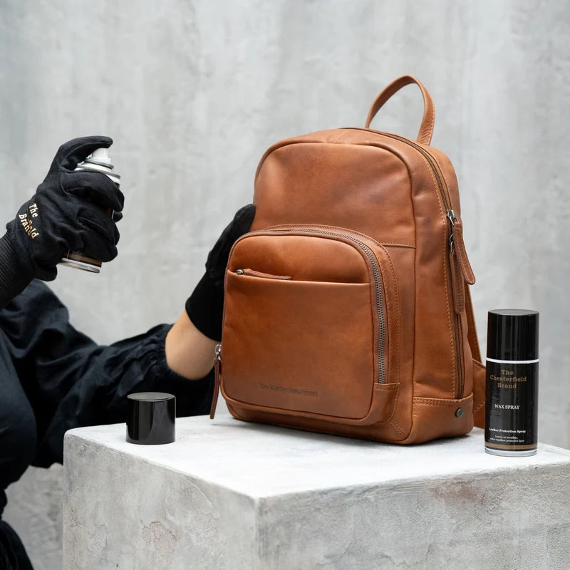 Lederwachs-Spray in Anwendung – Hand mit schwarzem Chesterfield-Handschuh sprüht auf cognacfarbenen Lederrucksack