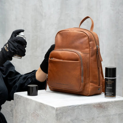 Lederwachs-Spray in Anwendung – Hand mit schwarzem Chesterfield-Handschuh sprüht auf cognacfarbenen Lederrucksack