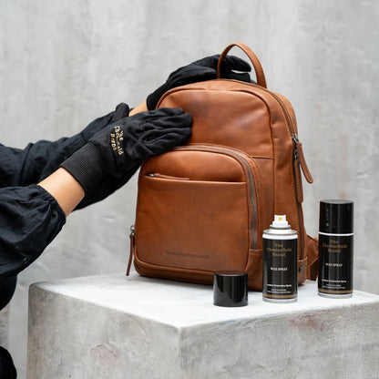 ederpflege mit Chesterfield Wax Spray – Hand mit Logo-Handschuh reibt cognacfarbenen Lederrucksack ein, zwei Sprühflaschen daneben